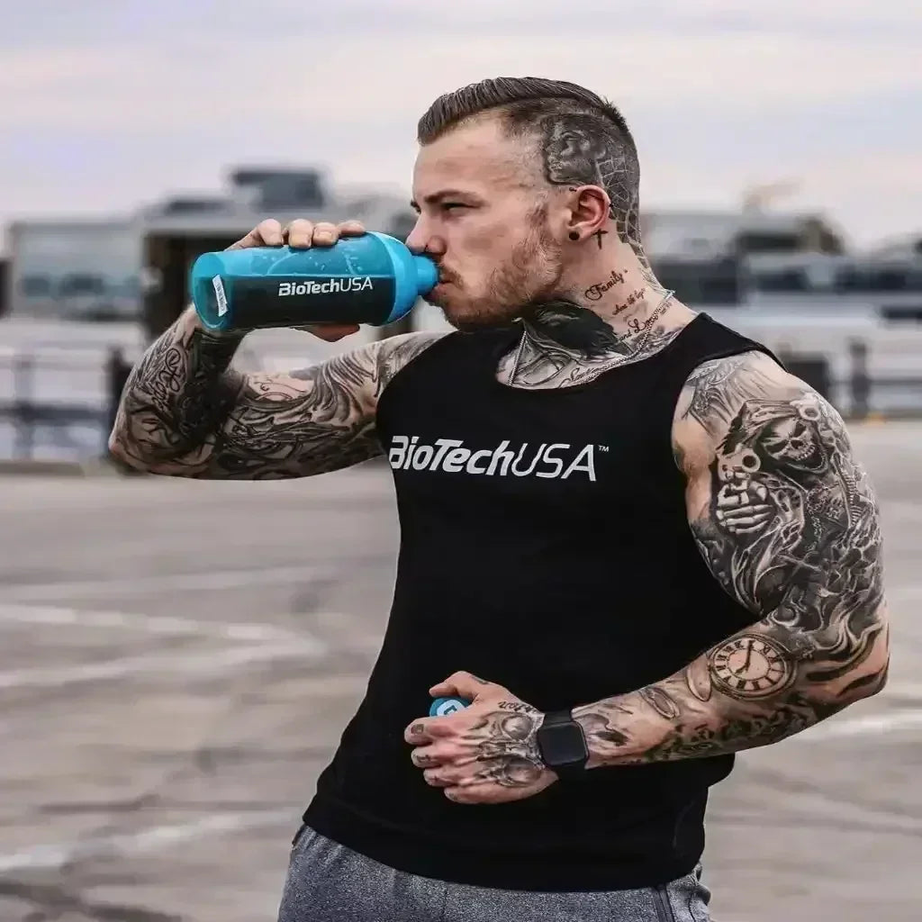 Muskulöser Mann mit Tattoos, BioTechUSA Tanktop und Shaker beim Trinken im Freien