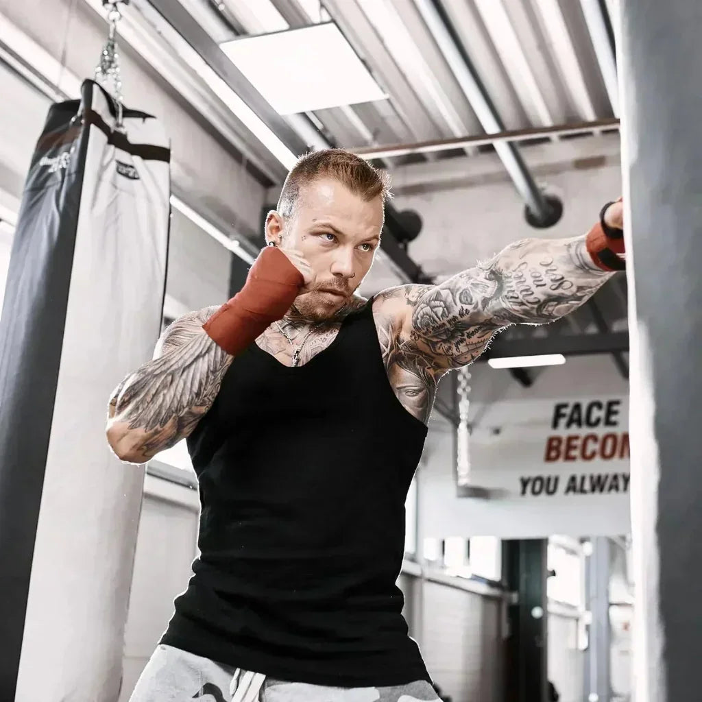Tattooierter Mann mit rotem Boxbandage trainiert an Boxsack im Fitnessstudio.
