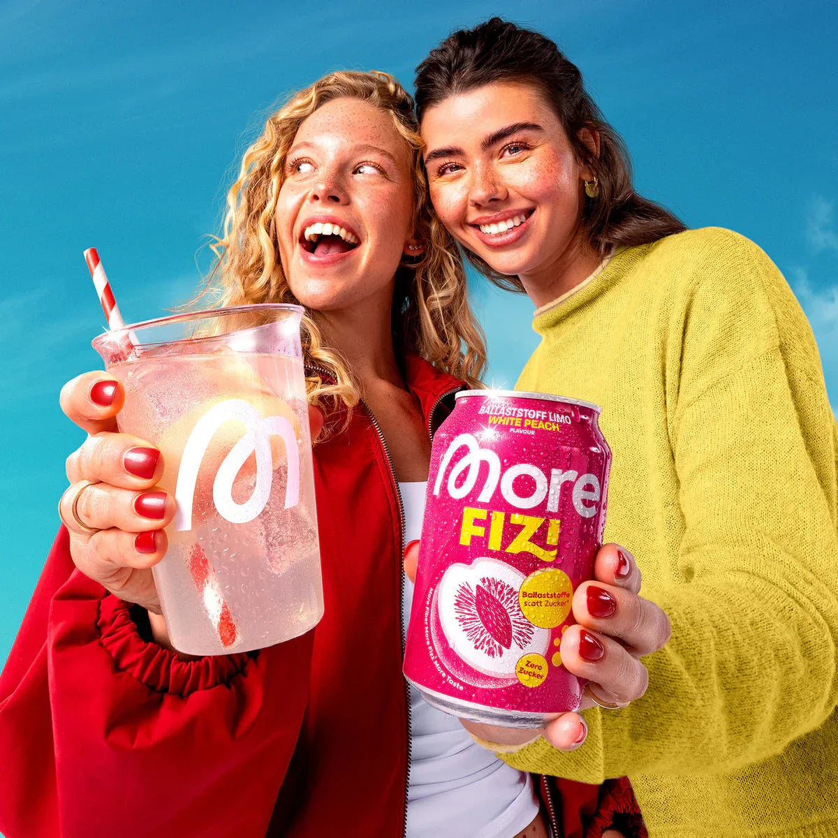 Zwei Frauen halten eine More FIZY Getränkedose und ein Glas mit Strohhalm vor blauem Himmel.